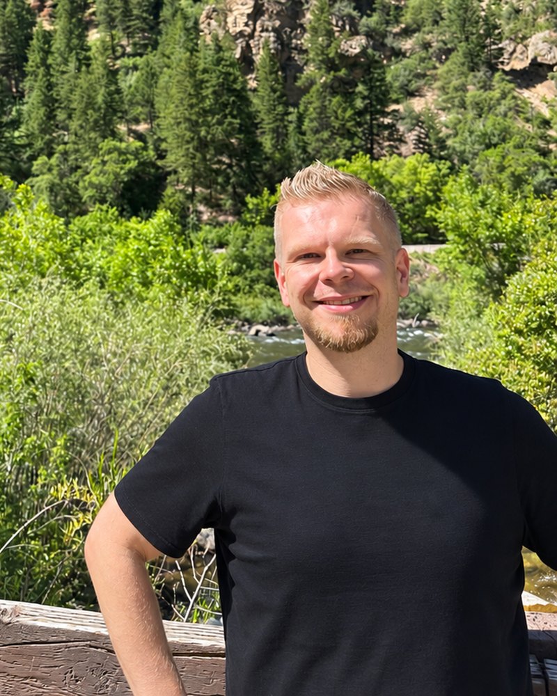 Tomasz Tomczyk outdoors, a pine-covered canyon and river behind him